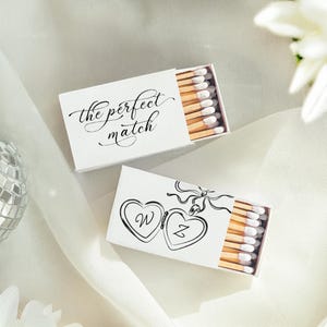 May include: Two white matchboxes with wooden matches. One box has the text "the perfect match" in elegant script. The other box features two connected hearts with the letters "W" and "Z" inside. The matches have white tips.
