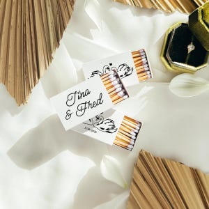 May include: Two white matchboxes featuring black ink illustrations of swans and the text Tina & Fred with the date 5/30/2026. The matchboxes are partially open, revealing wooden matches with white tips, arranged alongside a gold ring box.