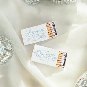 May include: Two rectangular matchboxes with white covers and light blue ink. One box features the names Lauren & Cole in script, while the other displays a graphic of two swans forming a heart shape with the text forever & ever below them.
