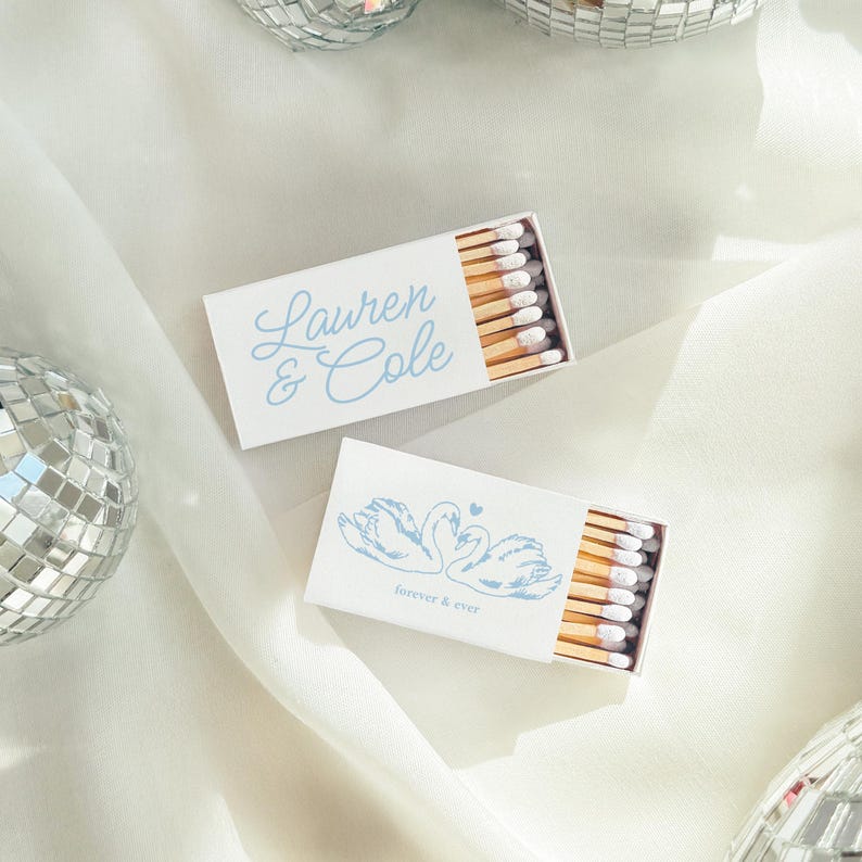May include: Two rectangular matchboxes with white covers and light blue ink. One box features the names Lauren & Cole in script, while the other displays a graphic of two swans forming a heart shape with the text forever & ever below them.