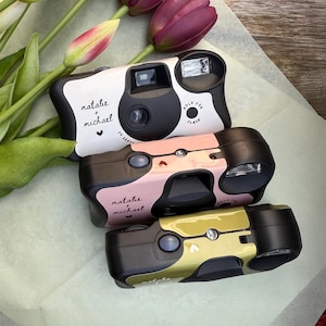 May include: Three disposable cameras with black plastic bodies and coloured front panels in white, light pink, and olive green. Each camera features the printed text "natalie + michael" and a small heart icon. The cameras include a flash unit.