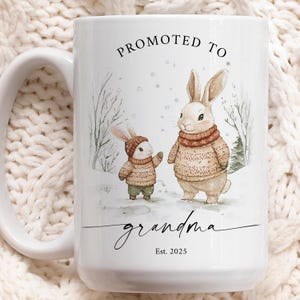 May include: White ceramic mug featuring a winter scene with two bunnies wearing sweaters. The text "Promoted to Grandma Est. 2025" is printed on the mug.