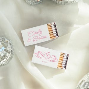 May include: Two rectangular white matchboxes with light pink text and graphics. One box features the names Cindy & Brian in script, while the other displays a line drawing of two swans forming a heart shape with the date 07-12-27 printed below.