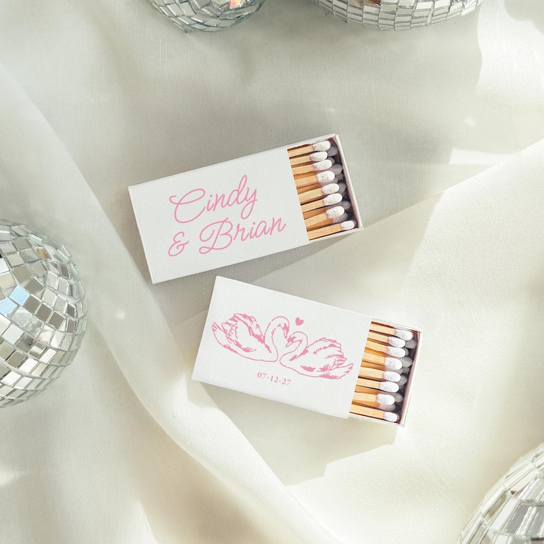 May include: Two rectangular white matchboxes with light pink text and graphics. One box features the names Cindy & Brian in script, while the other displays a line drawing of two swans forming a heart shape with the date 07-12-27 printed below.