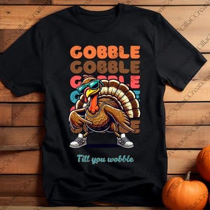 May include: Black t-shirt with a cartoon turkey wearing sunglasses and trainers. The shirt displays the words "GOBBLE GOBBLE GOBBLE" in orange, brown, and blue, with "Till you wobble" below. A fun Thanksgiving design.