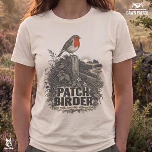 Pode incluir: Uma t-shirt de cor creme com um gráfico de um pisco-de-peito-ruivo pousado num poste de madeira entre folhagem. O texto diz PATCH BIRDER e same walk, every day, different list. O design inclui um nascer do sol sobre um campo ao fundo.