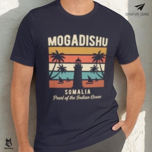 May include: A navy blue short-sleeve t-shirt featuring a graphic print of a lighthouse, palm trees, and sailboats against a sunset-colored striped background. The text reads MOGADISHU SOMALIA Pearl of the Indian Ocean with DEPARTURE LOUNGE.