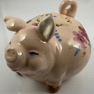 May include: A ceramic piggy bank in the shape of a pig, painted in a light pink hue. The pig features floral and vine designs in shades of purple, blue, and green. It has a slot on top for coins and a loop on the back.