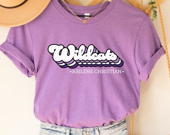 Może przedstawiać: Fioletowy T-shirt z napisem "Wildcats -ABILENE CHRISTIAN- " w retro stylu. Koszulka wisi na drewnianym wieszaku ze słomkowym kapeluszem i plecionym workiem.