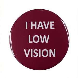 以下が含まれることがあります： 「I HAVE LOW VISION」の白い文字が書かれた、丸いバーガンディ色のバッジ。バッジは光沢があり、わずかな反射があります。テキストは中央に配置されており、明確な視覚的メッセージを提供します。