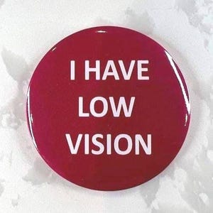 Puede incluir: Un botón redondo rojo con el texto blanco "I HAVE LOW VISION". El botón tiene un acabado brillante. El texto está centrado y es fácil de leer. El botón es probablemente de metal o plástico.