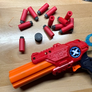 Peut inclure: Un pistolet jouet rouge et orange avec une poignée bleue et une cible noire et bleue. Le pistolet est entouré de fléchettes en mousse rouge.