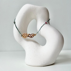 May include: A delicate bracelet featuring two small, carved wooden monstera leaf charms centered on a thin dark green cord. The cord is accented with tiny metallic silver beads and small, round, translucent green and red decorative beads.