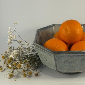 May include: A blue ceramic bowl with a geometric shape filled with orange citrus fruit. The bowl has a light blue glaze with a subtle pattern.