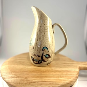 May include: A speckled ceramic pitcher with a curved spout and handle. The pitcher features a hand-painted design of two birds in teal and brown. The pitcher is sitting on a wooden cutting board.