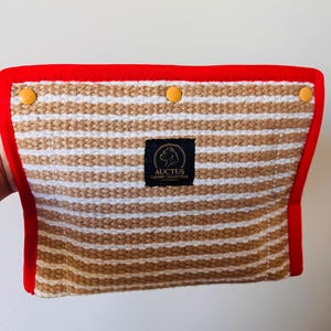 Könnte beinhalten: Eine rechteckige Tasche mit horizontalem beigefarbenem und weißem Streifenmuster und einem leuchtend roten Stoffrand. Drei gelbe Druckknöpfe sind entlang der Oberkante angebracht. Ein schwarzes Etikett in der Mitte zeigt den Text AUCTUS CANINE COLLECTIVE mit einem kreisförmigen Logo eines Hundekopfes.