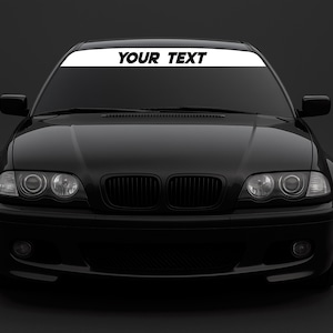 Puede incluir: Un coche negro con una pancarta blanca en la parte superior del parabrisas. La pancarta tiene las palabras "YOUR TEXT" en negro y en negrita. El coche tiene faros y una parrilla negra.