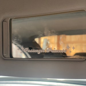 Puede incluir: Una visera para el sol de coche blanca con un espejo plateado y las palabras "Passenger Princess" en una fuente blanca cursiva.