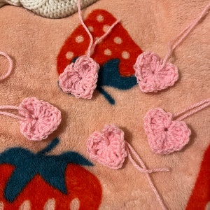 May include: Five pink crocheted hearts on a pink and red patterned fabric background.