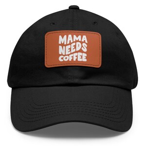 Op de afbeelding: Zwarte baseballpet met een bruin leren patch waarop in witte tekst "Mama Needs Coffee" staat.