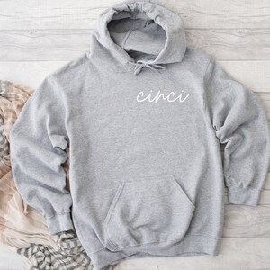 Puede incluir: Sudadera gris con capucha con la palabra "cinci" escrita en cursiva blanca en la parte delantera.