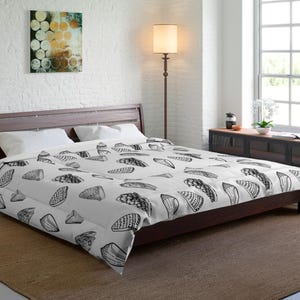 May include: A white comforter with a repeating pattern of black seashell illustrations. The bed is made up with white pillows and a dark brown wooden bed frame. The room has white brick walls and a brown rug.
