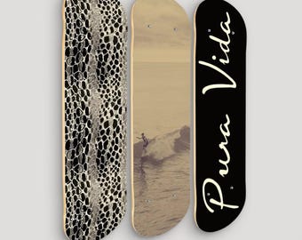 May include: Three skateboards hanging on a white wall. The skateboard on the left has a black and white snake skin pattern. The middle skateboard has a photo of a surfer riding a wave. The skateboard on the right is black with the text "Pura Vida" in white.