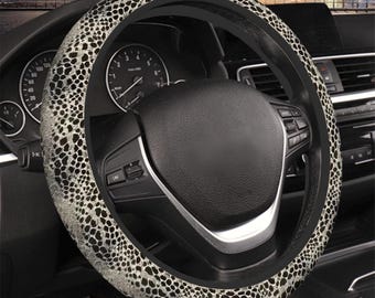 May include: A steering wheel cover with a black and white spotted pattern. The cover is wrapped around a black steering wheel in a car interior. The dashboard and other car features are visible in the background.