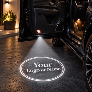 May include: A car door projector light installed in the underside of a vehicle door, casting a bright white circular beam onto the ground. The projection displays the text Your Logo or Name inside a double-lined circle, illuminating the dark pavement.
