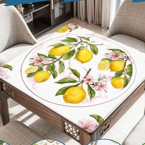 May include: A square wooden table with a white tablecloth featuring a lemon and floral design. The design includes yellow lemons, green leaves, and pink flowers. The table is surrounded by two beige chairs.