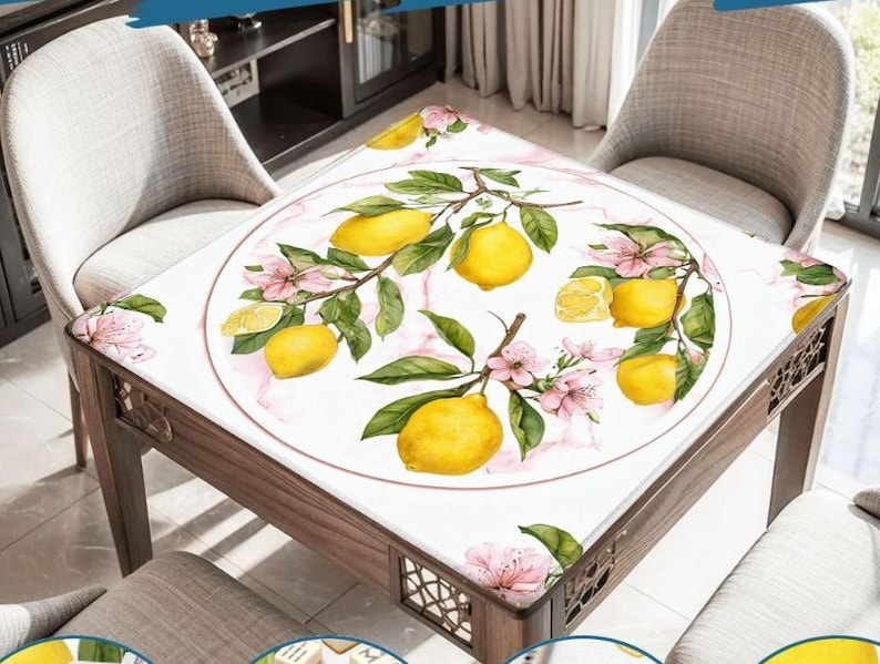 May include: A square wooden table with a white tablecloth featuring a lemon and floral design. The design includes yellow lemons, green leaves, and pink flowers. The table is surrounded by two beige chairs.