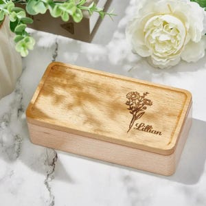 May include: A rectangular wooden box with a light wood finish. The lid features an engraved floral design and the name "Lillian" in a delicate script. The box is likely used for storing small items.