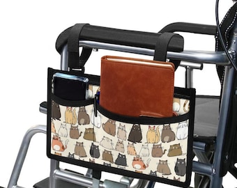 May include: A black wheelchair accessory bag with a white background and a pattern of cartoon cats. The bag has a zippered pocket on the front and two open pockets on the sides. A brown notebook is in the top pocket and a black phone is in the side pocket.