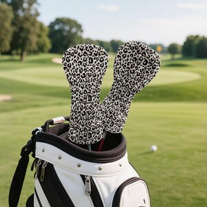May include: Two golf club head covers with a leopard print design, protruding from a white and black golf bag. The covers have a beige background with black spots. The golf bag is on a green golf course.