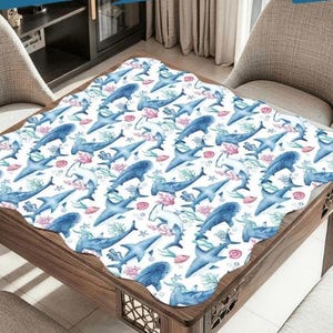 K&ouml;nnte beinhalten: Eine wei&szlig;e Tischdecke mit einem sich wiederholenden Muster aus blauen Haien, Korallen und Seegras. Die Tischdecke liegt auf einem Holztisch mit gewelltem Rand. Die Haie sind in verschiedenen Posen, was ein Unterwassermotiv ergibt.