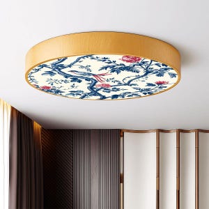 May include: A round flush mount ceiling light with a light wood frame. The light fixture features a cream-colored center with a blue and red floral and bird design. The light is mounted on a white ceiling.