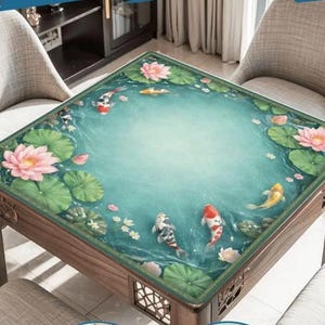 May include: Square wooden table with a glass top featuring a pond scene with koi fish, lily pads, and pink lotus flowers. The table has decorative carved details on the legs and frame. The table is surrounded by two chairs.