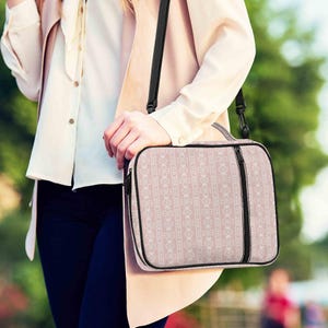 May include: A pink and white patterned shoulder bag with a black zipper and black strap. The bag has a rectangular shape and a handle on top. The bag is held by a person.