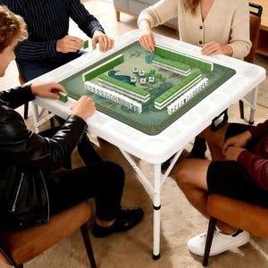 May include: A white folding Mahjong table with a green playing surface featuring a peacock design. The table is surrounded by players, with green and white tiles arranged on the surface. The table has built-in cup holders.