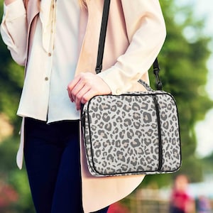 May include: A rectangular shoulder bag with a black strap and handle. The bag features a white background with a gray and tan leopard print pattern. The bag has a black zipper and trim.