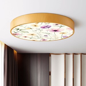 May include: A round flush mount ceiling light with a wooden frame. The light features a floral design with purple, pink, and yellow flowers on a cream background. The light fixture is a decorative element for home decor.
