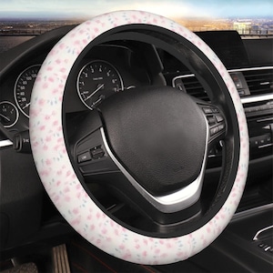 May include: A white steering wheel cover with a pattern of small pink flowers and green leaves. The cover is installed on a black steering wheel inside a car. The car's dashboard and other interior details are visible in the background.