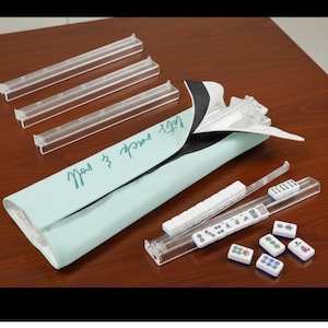 May include: A light blue Mahjong tile case with the text "Let's rack & roll" in cursive. The case is open, revealing clear plastic tile racks and white Mahjong tiles with various symbols and numbers. Several clear plastic racks are also visible.