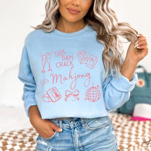 May include: Light blue sweatshirt with a pink Mahjong-themed graphic. The design includes champagne flutes, dice, tiles, a bow, and a disco ball. The text reads "Dot Bam Crack Mahjong".