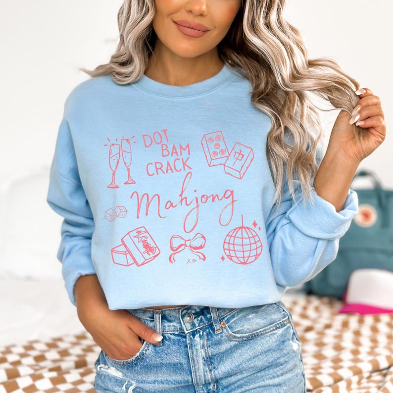 May include: Light blue sweatshirt with a pink Mahjong-themed graphic. The design includes champagne flutes, dice, tiles, a bow, and a disco ball. The text reads "Dot Bam Crack Mahjong".