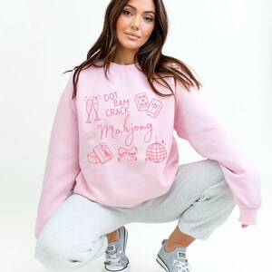 May include: A light pink sweatshirt with the words "Dot Bam Crack Mahjong" and related graphics in a matching pink color. The sweatshirt is paired with gray sweatpants and gray sneakers.