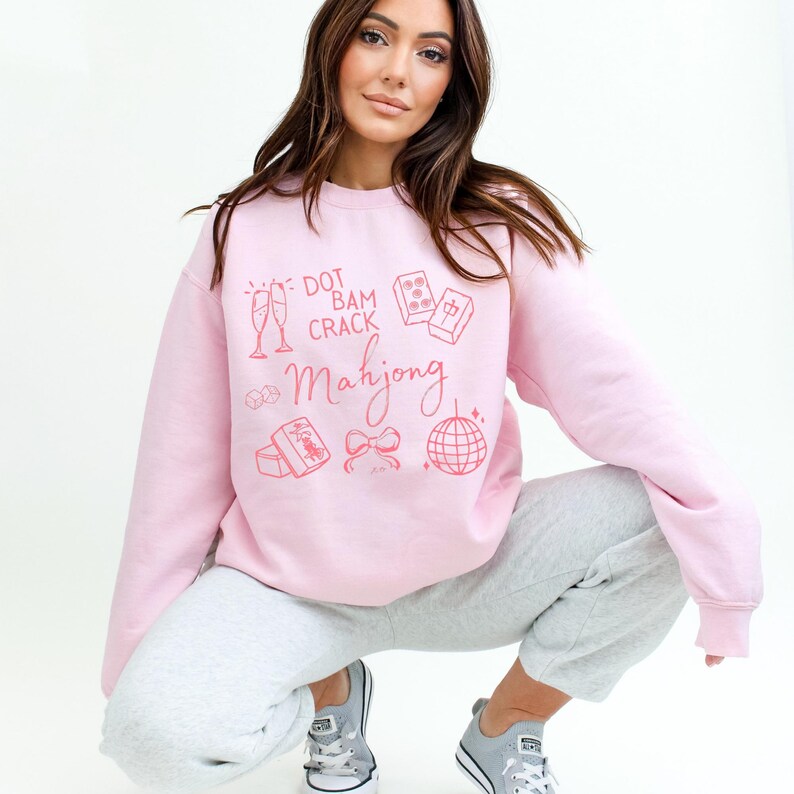 May include: A light pink sweatshirt with the words "Dot Bam Crack Mahjong" and related graphics in a matching pink color. The sweatshirt is paired with gray sweatpants and gray sneakers.