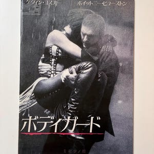 Può includere: Un poster cinematografico in bianco e nero con un uomo che porta una donna. Il titolo giapponese "Bodyguard" è visibile. Il poster include i nomi "Kevin Costner" e "Whitney Houston".