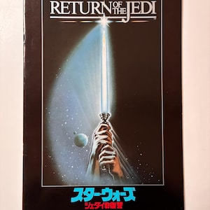 May include: A Star Wars: Return of the Jedi movie poster. The poster features a blue lightsaber held by a pair of hands, with a planet and stars in the background. The title is in white, with Japanese text below.