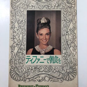 May include: Vintage promotional print for "Breakfast at Tiffany's" featuring a portrait of Audrey Hepburn. The image is framed with an ornate, grey and white floral design. The text "Breakfast at Tiffany's" is printed in green.